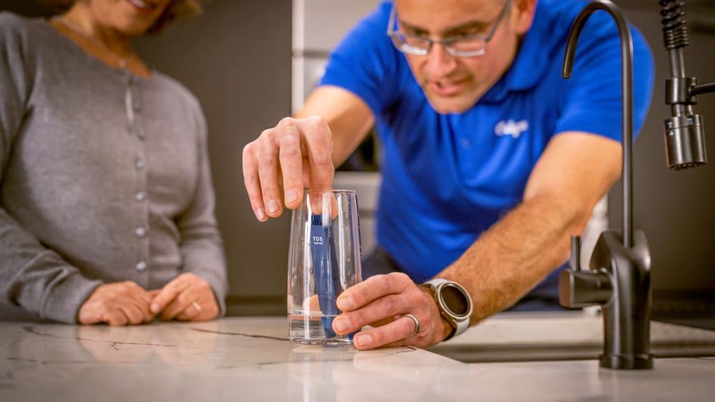 A man in a blue shirt uses a digital tester for water testing in a glass of water on a kitchen counter, while a woman in a grey sweater watches. A black sink faucet is visible in the foreground.