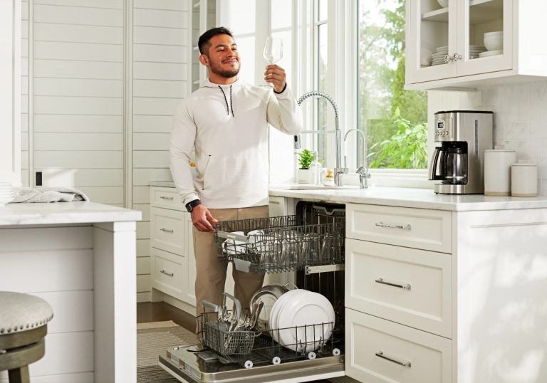 A man stands in a bright kitchen unloading a dishwasher. He smiles as he holds up a wine glass, admiring its sparkle, thanks to RO water systems. Clean dishes and utensils are visible in the open dishwasher racks.