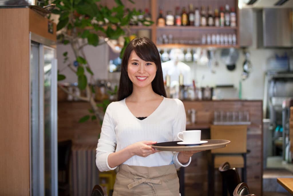 A woman wearing an apron smiles and holds a tray with a cup in a cozy café, where shelves of bottles, kitchen equipment, and commercial and industrial water treatment products are visible in the background.