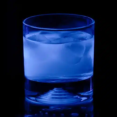 A rocks glass filled with a glowing blue liquid and ice cubes against a black background.