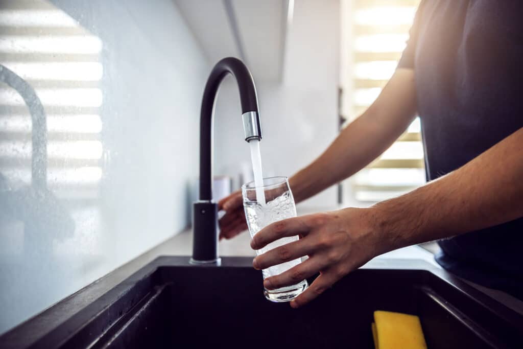 A person filling a clear glass with water from a kitchen faucet.