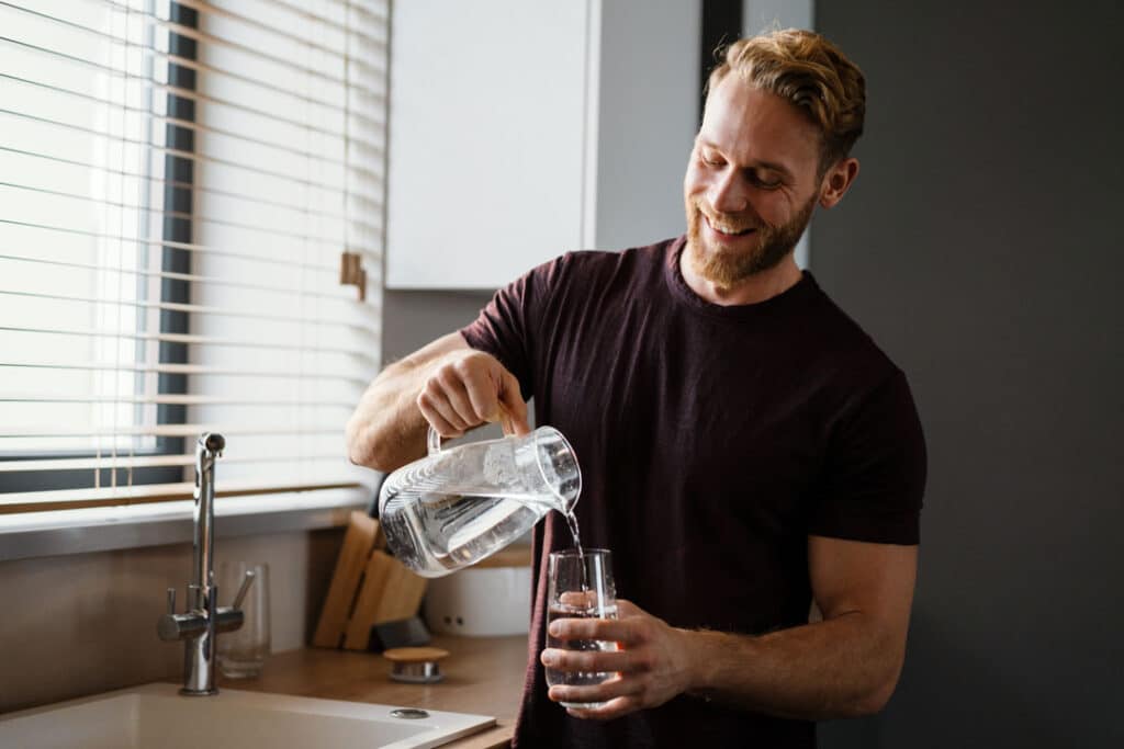 A smiling man pours water from a glass pitcher into a drinking glass in a kitchen.